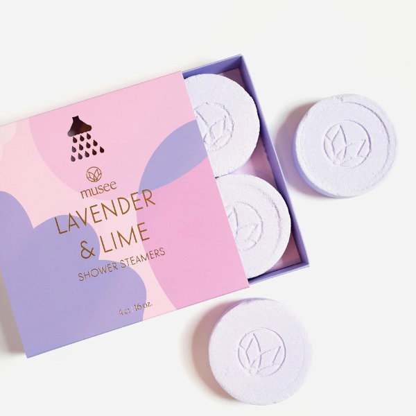 Commercial product photography for Musee featuring lavender and lime shower steamers on a white background, highlighting circular pale purple tablets with embossed lotus emblems and a decorative box with gold foil lettering and colorful abstract patterns
