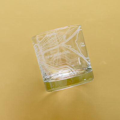 Etched glass rocks tumbler featuring a minimalist street map of New Orleans showing the French Quarter grid and Mississippi River with lowercase text against a solid mustard yellow background