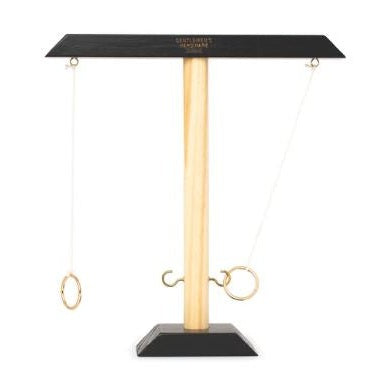 Wooden game by Gentlemen's Hardware featuring a tabletop hook and ring toss set with a light-grained central pillar, black trapezoidal base, and gold-toned metal rings suspended by strings against a plain white background