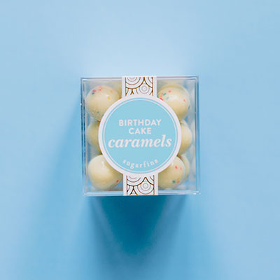 Commercial product photography by Sugarfina featuring a clear cube of Birthday Cake Caramels on a light blue background, showcasing off-white spherical candies with rainbow confetti specks and a blue circular label with gold scalloped details