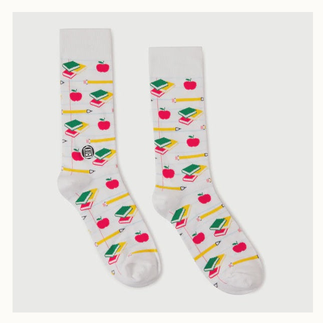 A pair of printed crew socks by BD showcasing a school-themed pattern on a neutral background, featuring vibrant red apples, yellow pencils, and stacks of green and red books over a faint notebook-lined design