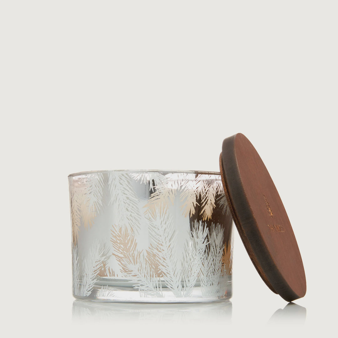 Thymes brand decorative glass candle jar featuring a metallic silver and white pine needle pattern in a neutral studio setting with a round wooden lid engraved with the brand logo propped against the side