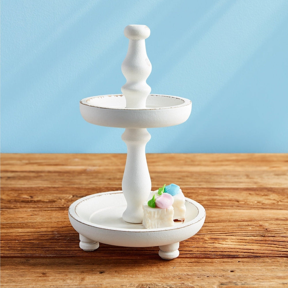 Studio photography of a two-tiered distressed white wooden tray on a rustic wood surface featuring miniature square cakes with pastel pink and blue frosting dollops against a solid light blue wall